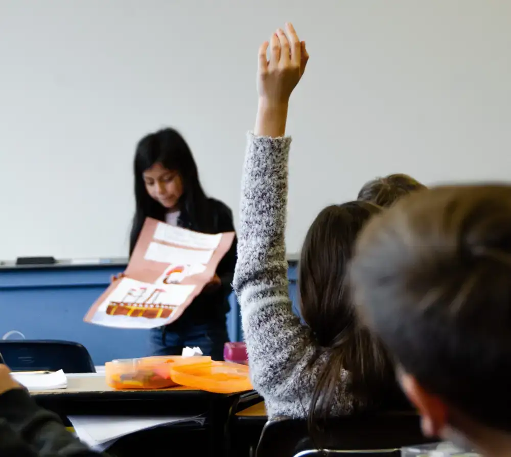 a young school girl doing a class presentation while another girl raises her hand to get her attention